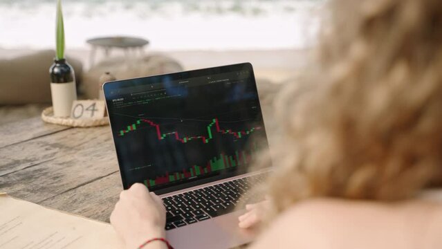 Female crypto trader at laptop checking candlestick charts online working remotely at outdoor tropical seaside cafe. Woman broker analyses exchange rates and bets on crypto stock by ocean closeup