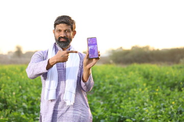 Happy rural Indian farmer showing his mobile phone towards the camera while looking at the camera, standing in a mustard field.