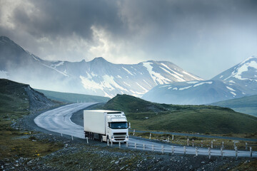 Large white transport truck transporting commercial cargo in semi trailer running on turning way highway road with scenic mountains mountaineous scenery in background. - 605555729