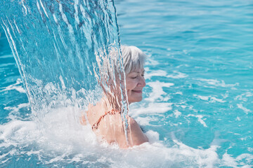 Senior caucasian smiling woman with gray hair enjoying falling on her shoulders flow of water. - obrazy, fototapety, plakaty