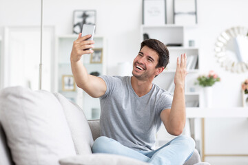 Young handsome happy man talking on a video call or takes a selfie on sofa in living room.