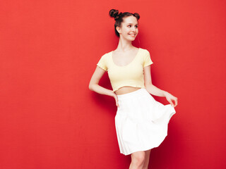 Young beautiful smiling female in trendy summer yellow t-shirt and skirt. Carefree woman with two horns hairstyle posing near red wall in studio. Positive model having fun. Cheerful and happy