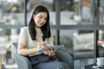 Attractive Asian young woman or businesswoman using tablet to search information on website for working and learning online. Business administration while sitting cross legged on sofa chair.