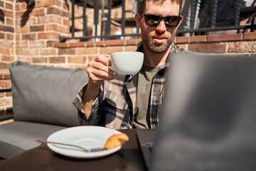 A businessman is sitting in a cafe with a cup of coffee and working on a sunny day. A handsome man looks at his laptop and drinks cappuccino.