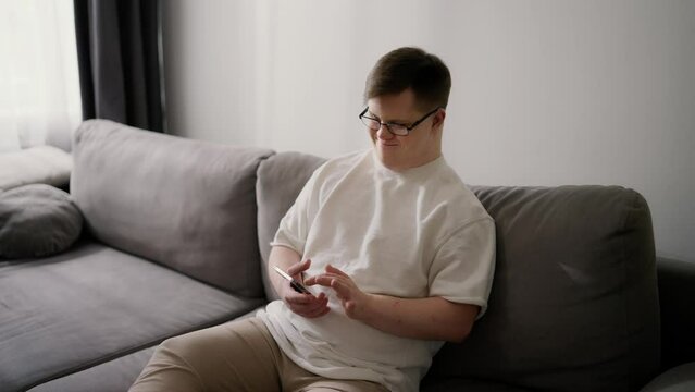 Smiling Downs Syndrome guy sitting on sofa using mobile phone, scrolling internet