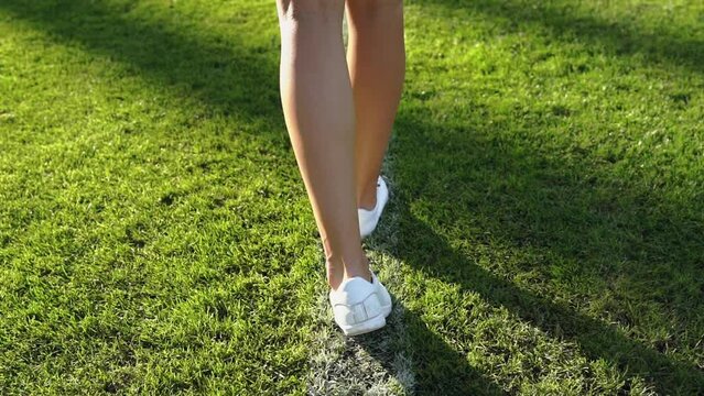 A caucasian girl in white sneakers walks along the white line of a football field close up. Back view portrait of a beautiful woman feet walking on the grass. Slow motion video footage in HD 25FPS