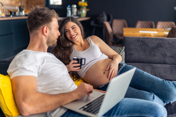 Expecting new parents are sitting on the couch planning the decor for the new children's room, the laptop is on his lap,she holds a cup of tea in her hand