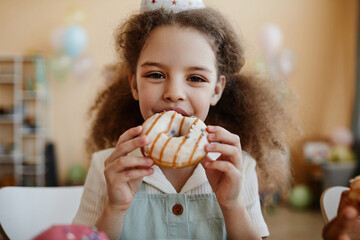 Front view portrait of cute little girl eating big donut during birthday party and looking at...