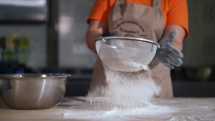 Unrecognizable female cook in gloves hitting sieve sifting powdered white flour in slow motion. Caucasian woman in apron preparing ingredient for baking in bakery kitchen indoors