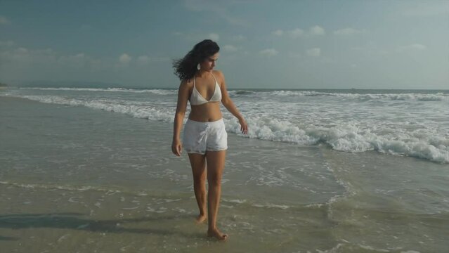 Woman in white bikini top and shorts plays and kicks water at beach, slow motion