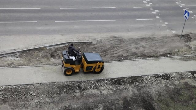 Video of road worker driver on mini roller compacts soil on pedestrian path under construction on side of autoroad. View from above