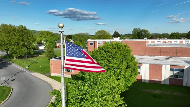 Rotational aerial shot of American flag waving in wind in front of public school building. Brick school, green grass and trees, blue sky. American flag, public high school theme.