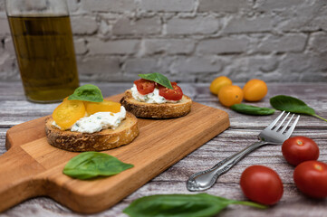 Tasty bruschetta with ricotta cheese, red and yellow cherry tomatoes and basil on a wooden cutting board. Ingredients on the background, image with bokeh effect.