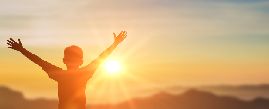 Boy raising hands over sunset sky, enjoying life and nature. Happy Kid in mountains looking on sun. Silhouette of child in sunlight rays. copy space
