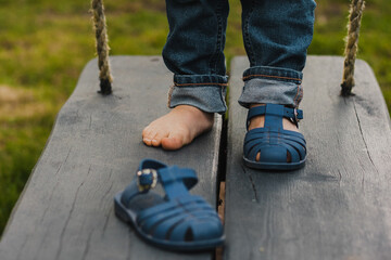 the boy's bare legs with blue sandals, one sandal taken off. a bare child's leg on a dark wooden swing in blue jeans.
