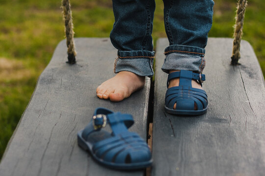 the boy's bare legs with blue sandals, one sandal taken off. a bare child's leg on a dark wooden swing in blue jeans.