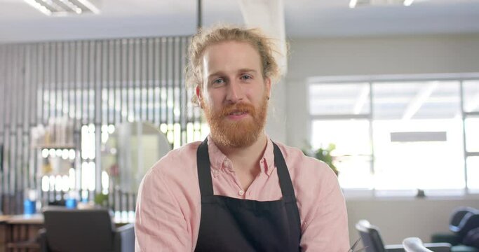 Portrait of bearded caucasian male hairdresser in apron smiling at hair salon, slow motion