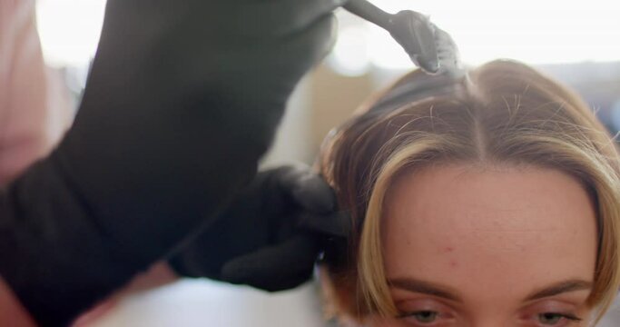 Gloved hand of caucasian hairdresser dyeing female client's hair with brush at salon, in slow motion