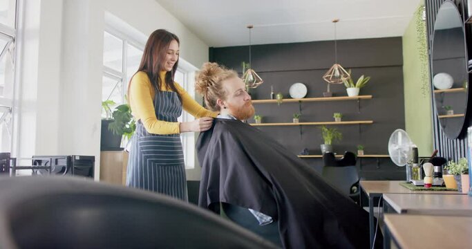 Happy caucasian female hairdresser putting cape on male client at hair salon, in slow motion