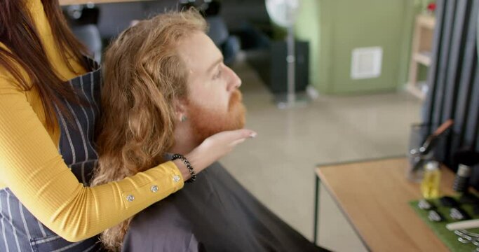 Caucasian female hairdresser touching beard of male client and advising at salon, in slow motion