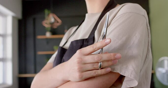 Midsection of caucasian female hairdresser in black apron holding scissors, in slow motion