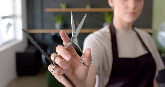 Midsection of caucasian female hairdresser in black apron holding scissors and comb, in slow motion