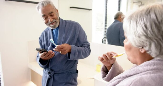 Happy senior biracial couple brushing teeth,using smartphone in bathroom, unaltered, in slow motion