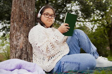 Fototapeta premium Happy woman with Down syndrome reading and listening to music in the park