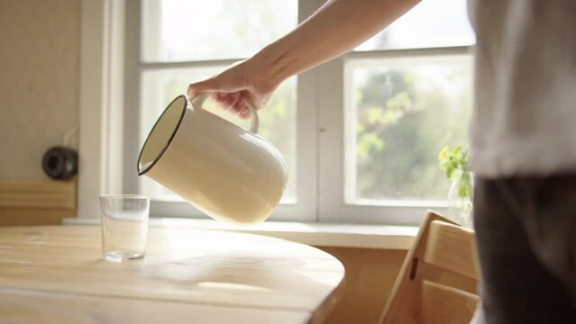 Person pours fermented milk kefir into glass on table, backlit by window