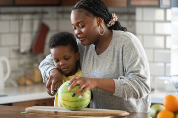 Fototapeta premium Cheerful African-American mother and son in the kitchen preparing a healthy fruit snack
