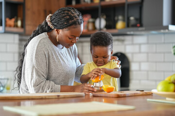 Cheerful African-American mother and son squeezing an orange to make orange juice. Son helps mother...