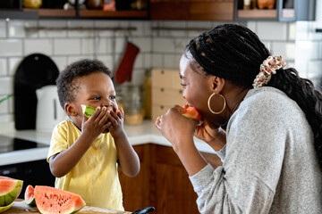 Cheerful African-American mother and son eating a tasty watermelon at home. Flavors of summer - 605613164
