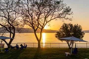 Silhouette of trees and people by the lake with a golden sky at sunset. Beautiful dramatic orange sky landscape near the Sea of Galilee, Israel.