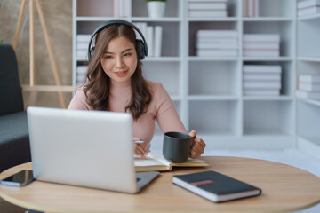 Portrait of a teenage Asian woman using a laptop computer, wearing headphones and using a notebook to study online via video conferencing and holding coffee cup on a wooden desk in living room.