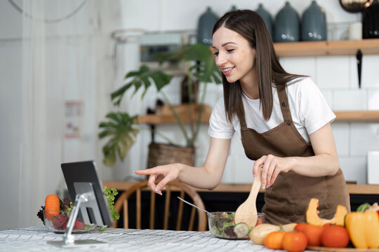 Image of young pretty lady in kitchen and cooking the salad. Looking at tablet computer