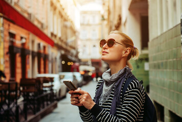 A young adult blonde female traveler walks and smiles around a European city, using a smartphone, taking photos and studying information from travel apps