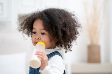 Cute curly hair biracial little girl sucking milk from bottle. Hungry kid holding milk bottle happy...