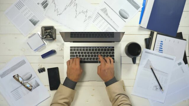 Male arms of businessman typing text on laptop keyboard at white desk. Top view hands of manager working on notebook in office. Diagrams with statistics at background. Concept of business lifestyle