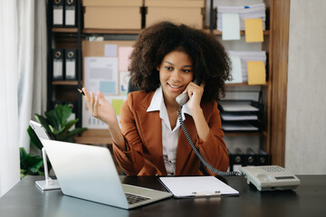 Obraz premium Young African woman typing on tablet and laptop while sitting at the working wooden table modern office
