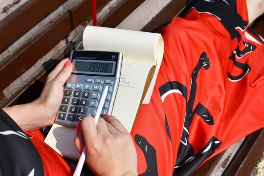 Closeup shot of a woman sitting on bench using calculator to write on a notebook