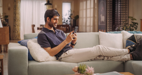 Handsome Indian Man Lying on Sofa Using Smartphone, Exploring Online Shopping, Remote Work, And Watching funny Videos on Social Media From The Comfort Of Cozy Home.