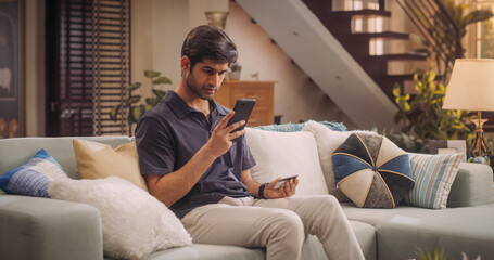 Handsome Indian Man Using Credit Card and Smartphone for Online Shopping at Home. Smiling Happy Guy Purchasing Good at e-Store. e-Commerse Concept. Medium Shot