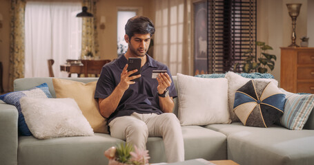 Handsome Indian Man Using Credit Card and Smartphone for Online Shopping at Home. Smiling Happy Guy Purchasing Good at e-Store. e-Commerse Concept. Medium Shot