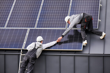 Installers building photovoltaic solar module station on roof of house. Men electricians in helmets installing solar panel system outdoors. Concept of alternative and renewable energy.
