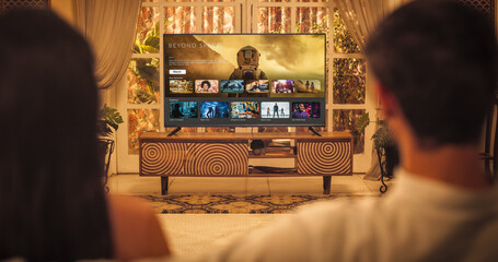 Back View of Young Indian Couple Watching TV while Sitting on a Couch in the Living Room. Relaxed Evening for Enjoying Favourite Movies, TV Shows, Content and Ads Thank to Streaming Online Service