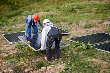 Workers building solar panel system. Men technicians in helmets carrying photovoltaic solar module outdoors. Concept of alternative and renewable energy.