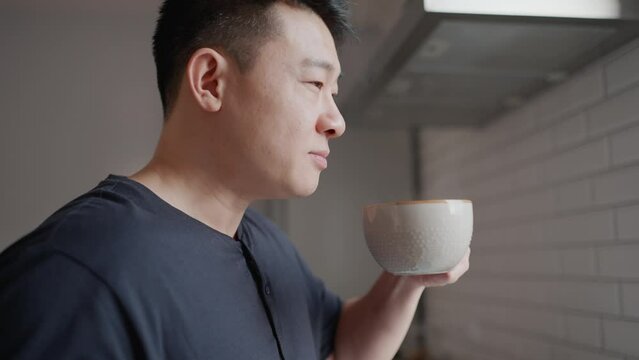 Smiling asian man drinking cup of coffee in kitchen at home