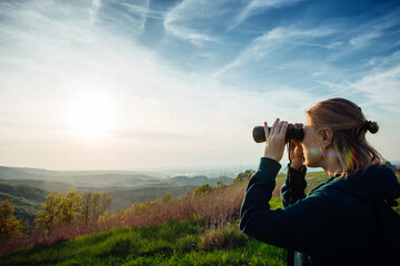 A young adult female hiker and traveler looks through binoculars in the mountains in the magical evening light of a sunset