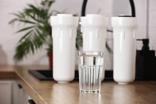 Glass of clean drinkable water and set of filter cartridges on wooden table top in a kitchen, houseplant. Installation of reverse osmosis water purification system. Concept household filtration system