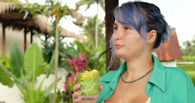 Half-turn portrait of pretty woman drinking green cool soft drink from glass and smile, looking at distance, have good time at trendy Balinese cafe. Young woman have blue colored hair